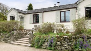 A two-story house with a stone staircase leading to the entrance, surrounded by lush greenery and purple flowers. - 1 Bed in Llanerchymedd (oc-wai212) (Carmel)