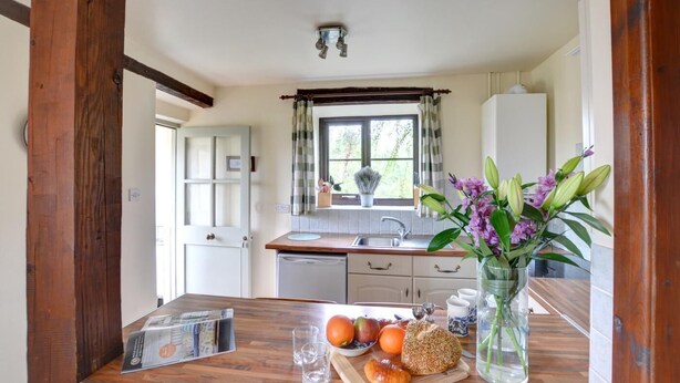 A kitchen with a wooden table, a window with curtains, and a vase with flowers. - 1 Bed in Llanerchymedd (oc-wai212) (Carmel)
