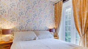 Een slaapkamer met bloemen behang, een bed met witte lakens, twee bedlampen en een raam met gordijnen.