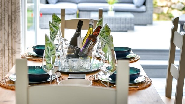Una mesa de comedor dispuesta con una botella de champán, copas y platos, con vistas a un sofá y follaje al fondo.