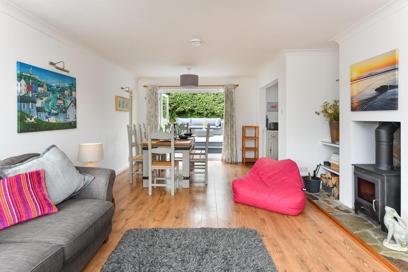 Een ruime woonkamer met een grijze bank, een rode poef, een eettafel met stoelen, een open haard en uitzicht op de omgeving.