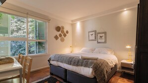 Een slaapkamer met een bed, nachtkastje, lamp en uitzicht op bomen door het raam.