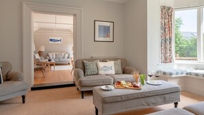 A living room with a sofa, a dining area, and a window with curtains. - 7 Bed in Aldeburgh (oc-gbl) (Aldeburgh)