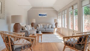 A spacious living room with a sofa, two armchairs, a coffee table, and a dining table. There are two lamps, a framed picture on the wall, and large windows with a view of greenery outside. - 7 Bed in Aldeburgh (oc-gbl) (Aldeburgh)