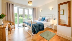 Une chambre à coucher comprenant un lit, des tables de chevet, un miroir, une fenêtre avec des rideaux, un plancher en bois et une vue sur la verdure à l’extérieur.