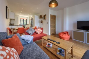 Un salon moderne avec un canapé, une table basse et un téléviseur. On aperçoit, au fond, un coin cuisine avec une table à manger et des chaises.