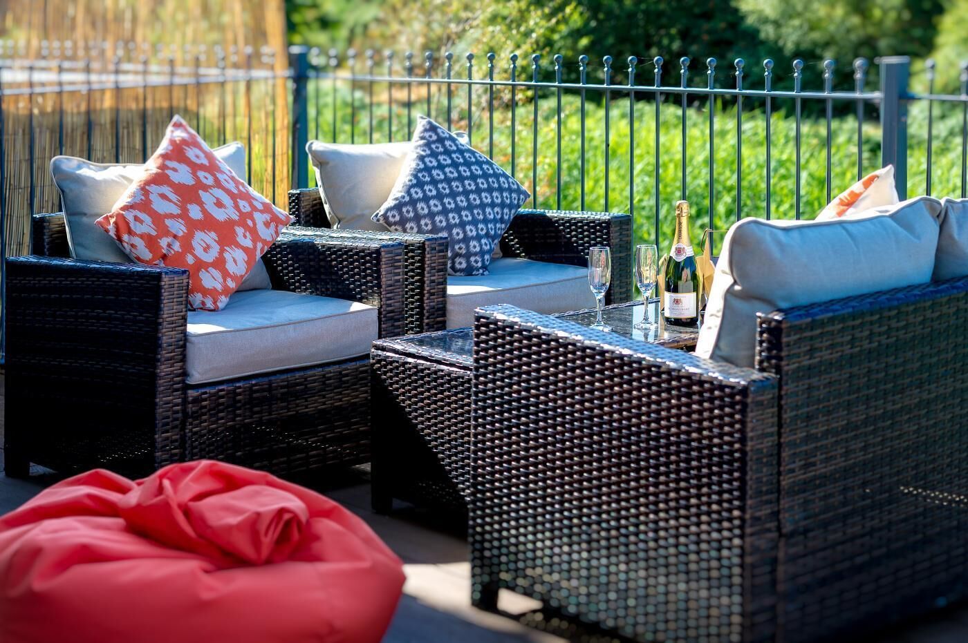 Mobilier d’extérieur en osier, agrémenté de coussins, une table en verre avec une bouteille et des verres, et un pouf rouge.