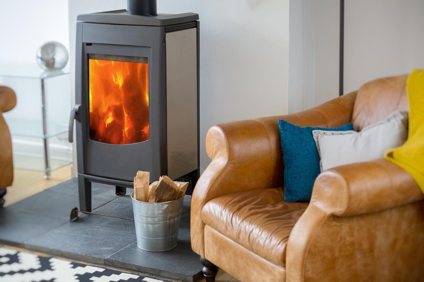 A modern living room with a black wood-burning stove, a leather sofa with cushions, and a metal bucket filled with firewood.