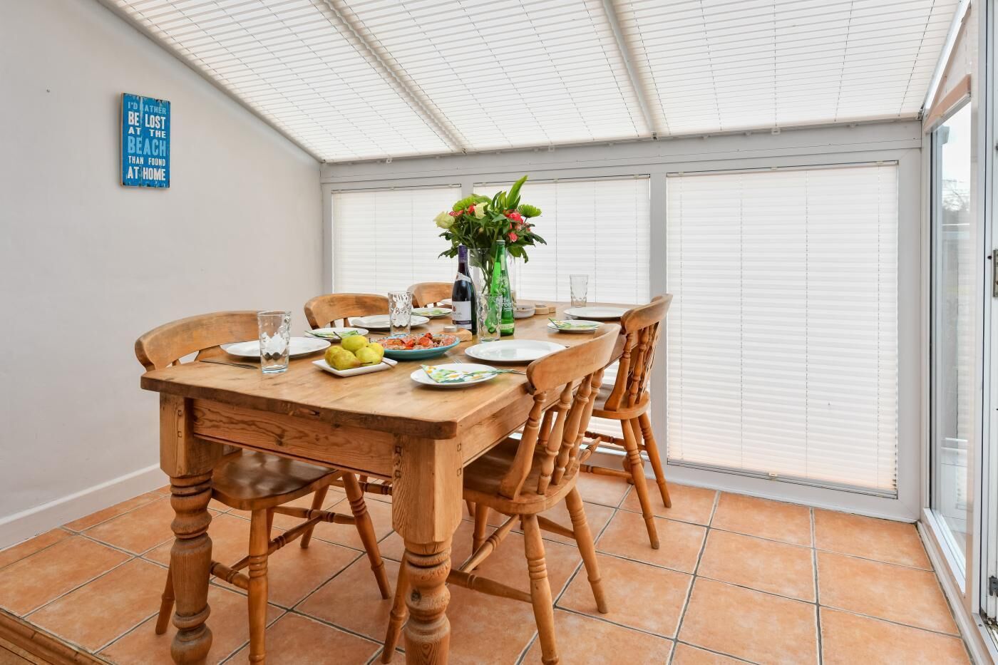 Uma área de refeições com mesa de madeira, cadeiras e uma mesa com tampo de vidro. Há um vaso com flores, pratos com comida e uma garrafa de vinho.
