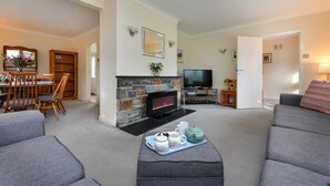 A living room with a stone fireplace, grey sofas, a dining area with wooden furniture, and a television. - 4 Bed in Llanbedrog (oc-sunnyd) (Llanbedrog)