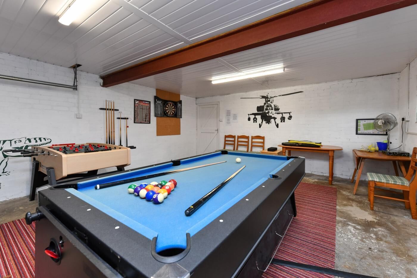 Une table de billard avec des billes, une table de baby-foot et un jeu de fléchettes dans une pièce aux murs blancs et au plafond en bois.