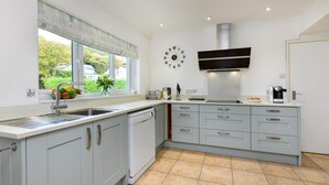 A modern kitchen with grey cabinets, a built-in dishwasher, a large window with a view of greenery, and a wall-mounted clock. - 4 Bed in Llanbedrog (oc-sunnyd) (Llanbedrog)