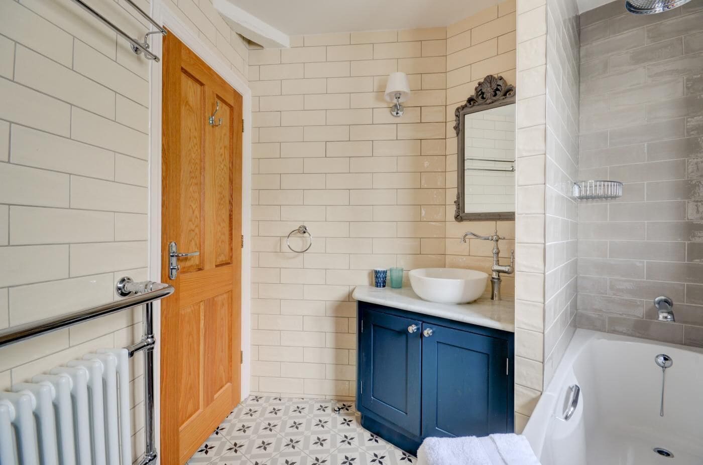 A bathroom with a wooden door, white tiled walls, a blue cabinet, a white sink, a mirror, and a bathtub.