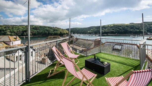 Een terras met lounge stoelen in rood en wit gestreept patroon, een tafeltje met een fles, en uitzicht op een jachthaven met boten.