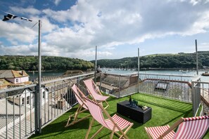 A terrace with red and white striped lounge chairs, a small table with a bottle, and a view of a marina with boats. - 3 Bed in Fowey (oc-p00647) (Fowey)