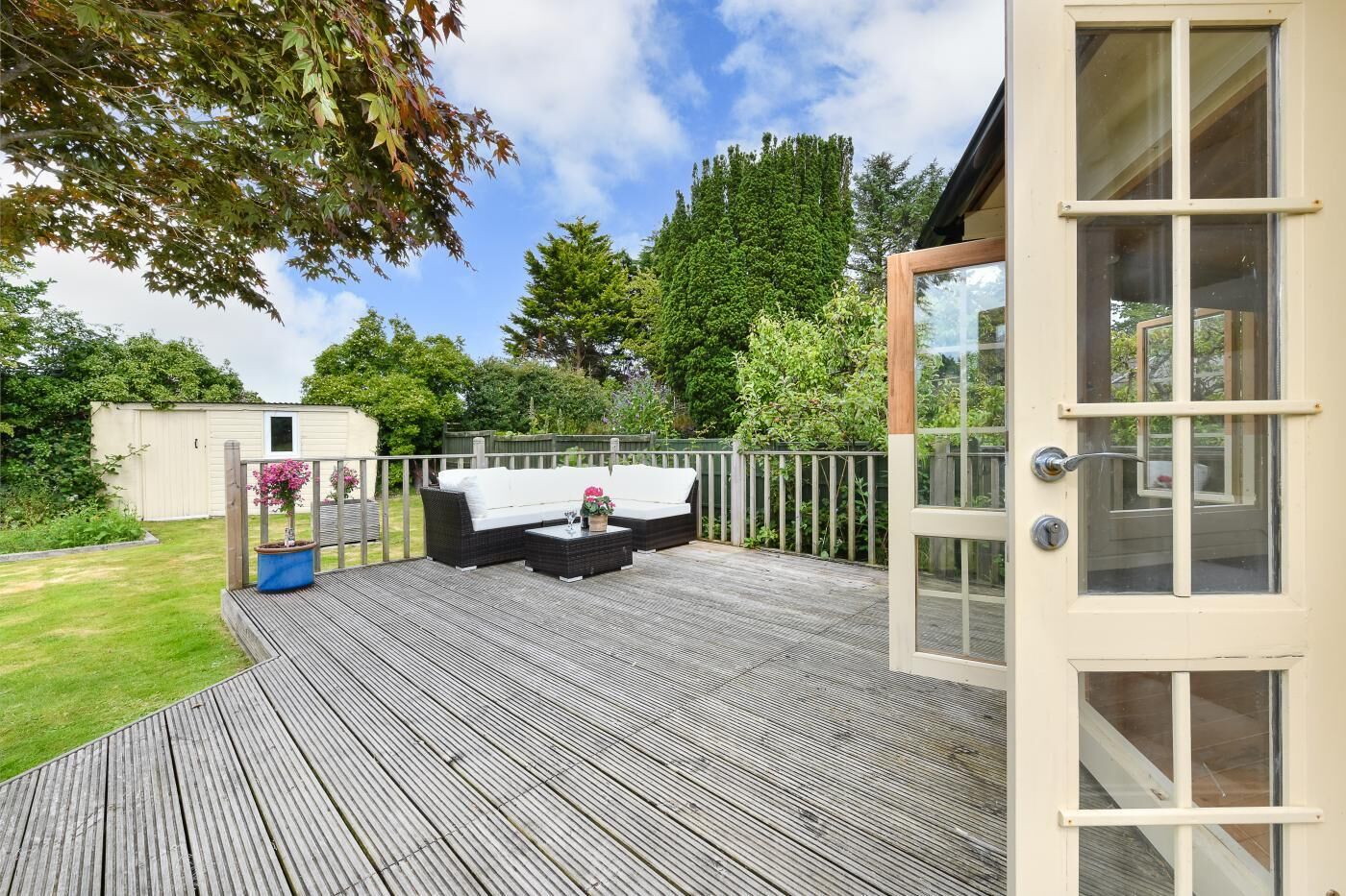 Une terrasse extérieure avec un canapé et une table basse, une terrasse en bois, une porte blanche et un jardin avec des arbres et un abri de jardin.