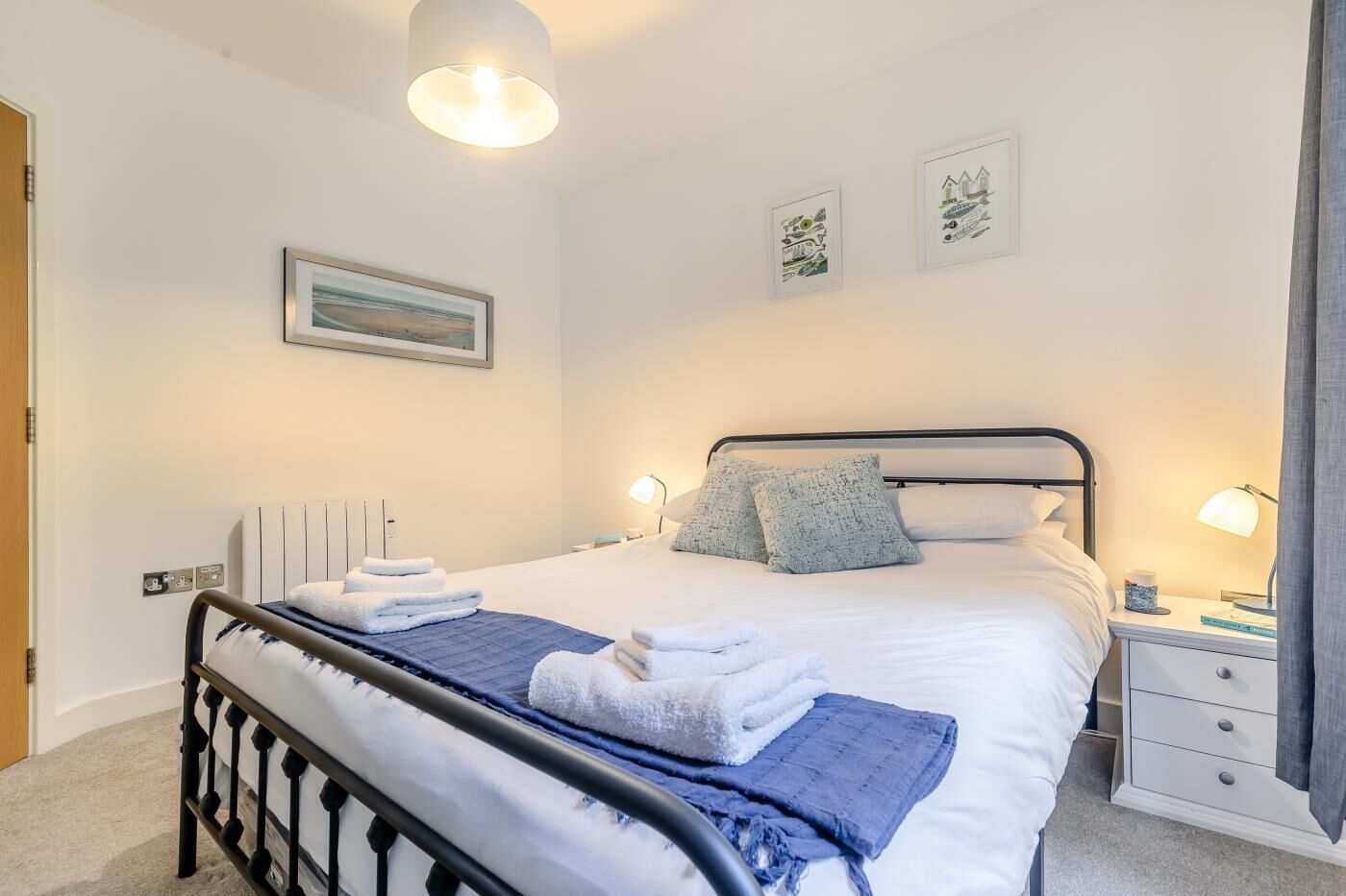 Un lit bien fait, avec du linge de lit blanc et une couverture bleue, une table de chevet avec une lampe, et un radiateur fixé au mur.
