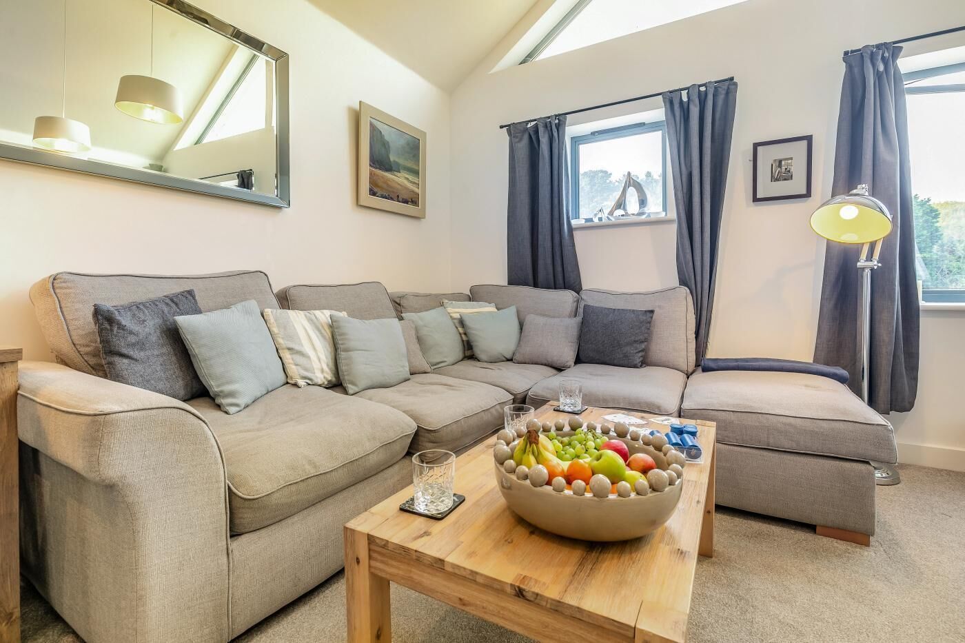 A modern living room with a grey sectional sofa, a wooden coffee table with fruit, and a large window with curtains.