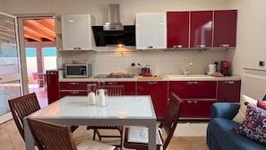A compact kitchen with red and white cabinets, a microwave, a toaster, and a coffee maker. There is a dining table with chairs and a sofa with a patterned cushion.