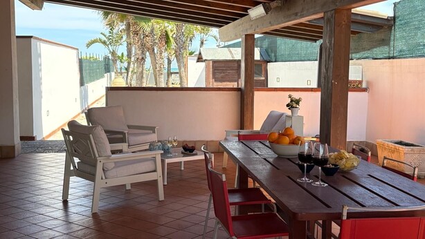 Un patio techado con una mesa de madera lista para comer, sillas blancas acolchadas y vistas a palmeras y edificios al fondo.