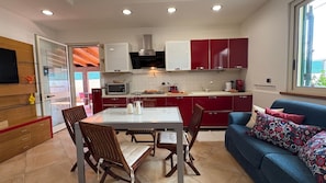 A compact kitchen with red cabinets, a dining table with chairs, a blue sofa with patterned cushions, and a window with a view of the outside.