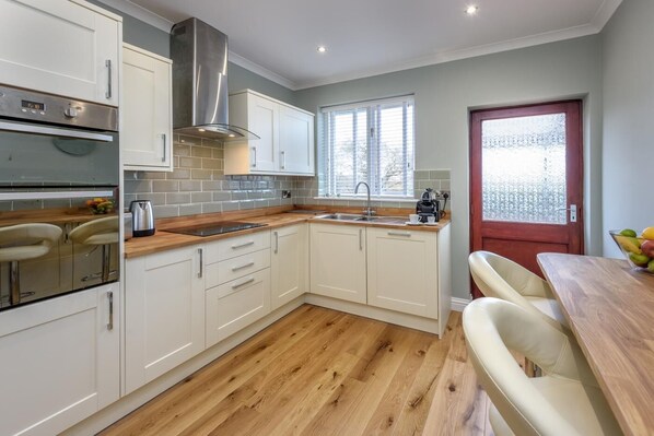 A modern kitchen with white cabinets, a stainless steel range hood, a wooden countertop, and a red door. - 3 Bed in Tudweiliog (oc-1brodw) (Tudweiliog)