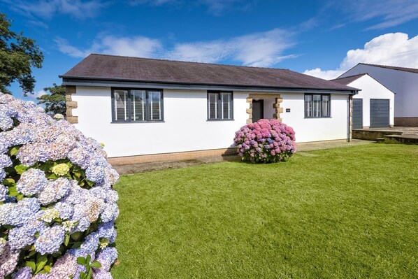 A two-story house with a white exterior, a dark roof, and a front door. There is a well-maintained lawn and a bush with pink flowers in front of the house. - 3 Bed in Tudweiliog (oc-1brodw) (Tudweiliog)