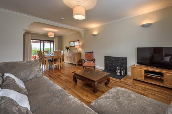 A spacious living room with a wooden floor, a large sofa, a dining table with chairs, a fireplace, and a television. - 3 Bed in Tudweiliog (oc-1brodw) (Tudweiliog)