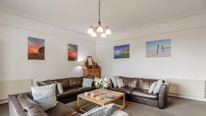 A living room with a brown leather sectional sofa, a wooden coffee table, and a chandelier. There are three framed landscape paintings on the walls. - 3 Bed in Llanbedrog (oc-thehol) (Llanbedrog)