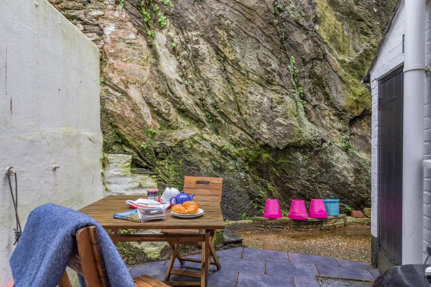 A wooden table with a plate of food, a book, and a teapot in a rustic outdoor setting with a stone wall and a wooden bench.