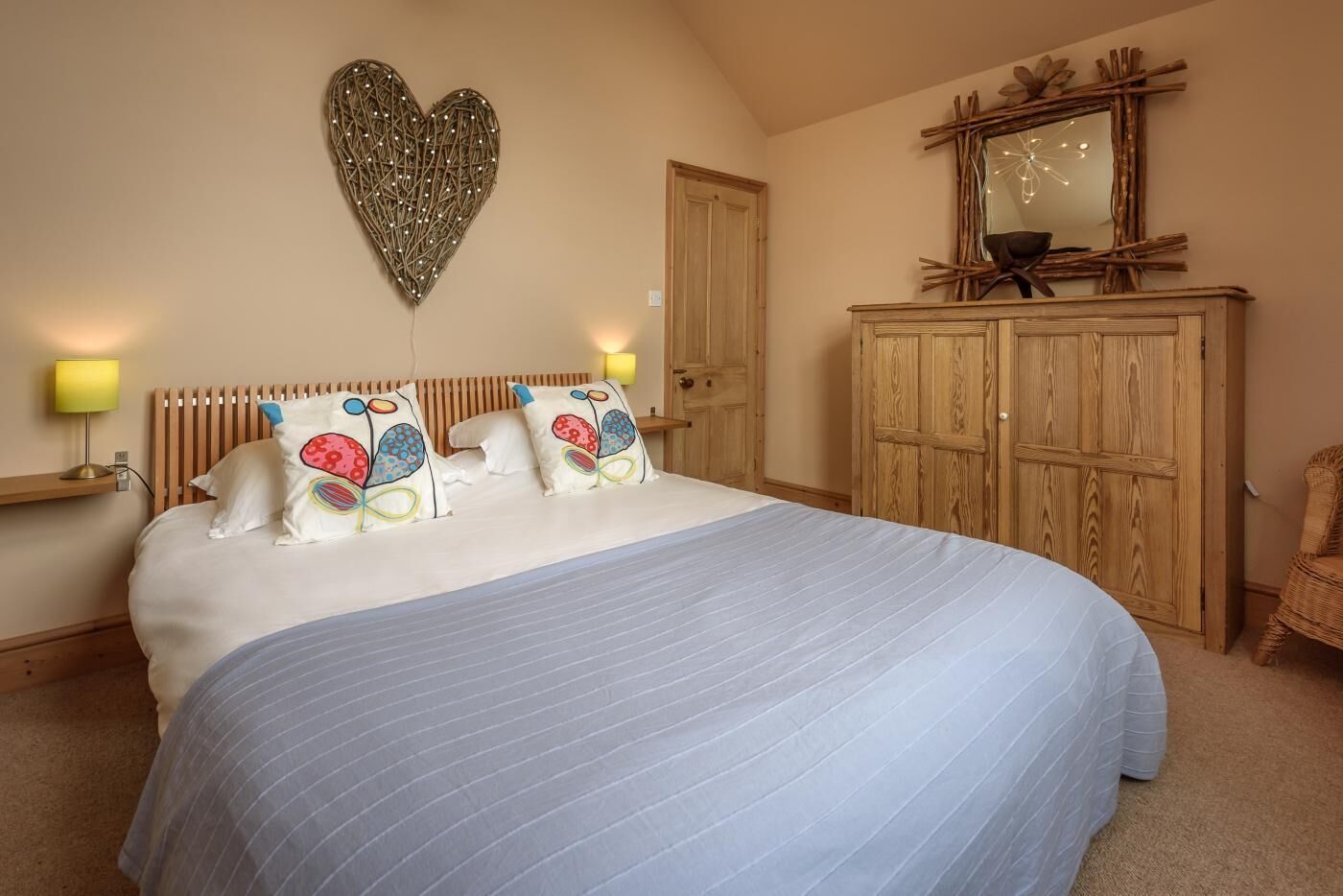 A bedroom with a large bed, wooden headboard, two bedside lamps, a heart-shaped wall decoration, a wooden wardrobe, and a mirror with a rustic frame.