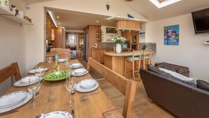 A dining area with a wooden table set for four, a kitchen in the background, and a TV mounted on the wall. - 4 Bed in Mynytho (oc-angmyn) (Mynytho)