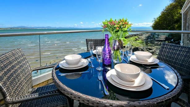 A round table with white plates, bowls, and cutlery, set against a scenic waterfront view. - 2 Bed in Llanbedrog (oc-2thebo) (Llanbedrog)