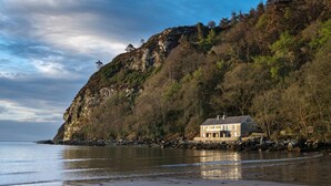 A coastal house with a stone exterior, situated on a sandy beach with a rocky cliff in the background. - 2 Bed in Llanbedrog (oc-2thebo) (Llanbedrog)