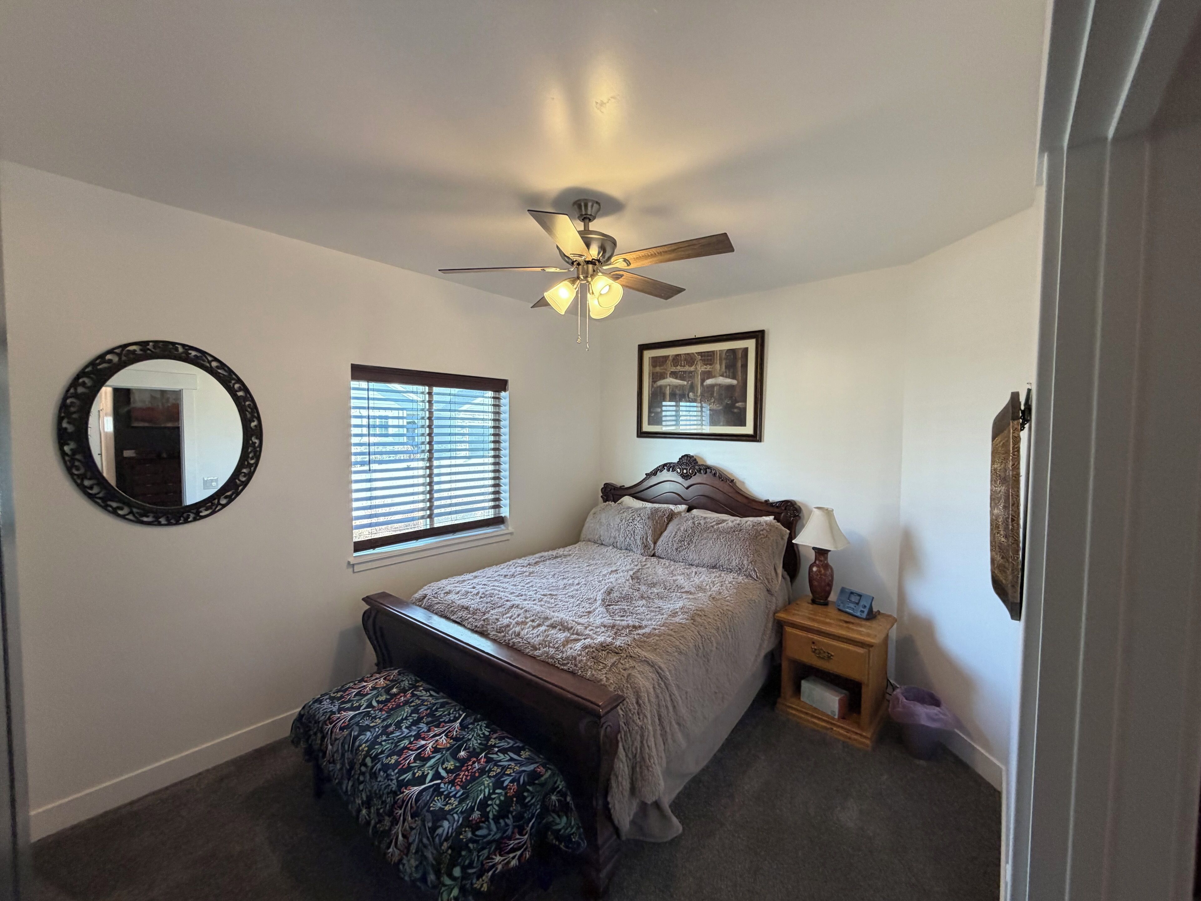 Un dormitorio con cama, ventilador de techo, ventana con persianas, mesita de noche con lámpara, espejo y un cuadro en la pared.