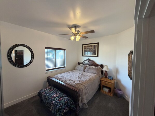 A bedroom with a bed, a ceiling fan, a window with blinds, a bedside table with a lamp, a mirror, and a framed picture on the wall. - Cozy & Comfy Mountain-View Home Airport 15 min (West Valley City)