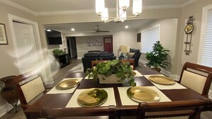 A dining area with a wooden table set for four, surrounded by chairs. There is a chandelier above the table, and a television is mounted on the wall in the adjacent living room. - Peaceful 3BR NW Raleigh Ranch Home. Pet friendly, RDU, UNC/Rex, Crabtree Mall (Raleigh)