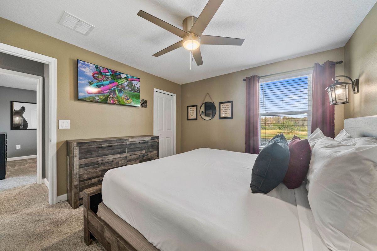 A bedroom with a bed, a ceiling fan, a window with curtains, and a wall-mounted TV.
