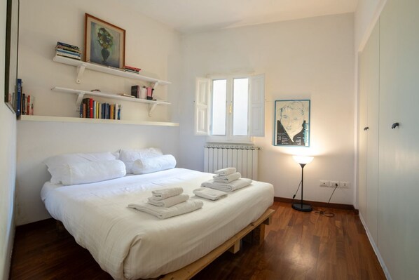 A bedroom with a wooden bed, white bedding, a radiator, a standing lamp, and shelves with books and a framed picture. - San Niccolò Elegant Apartment (Florence)