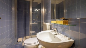 Ett badrum med en dusch som är avskärmad med glasväggar, en vit toalett och en vit handfat med en glashylla ovanför.