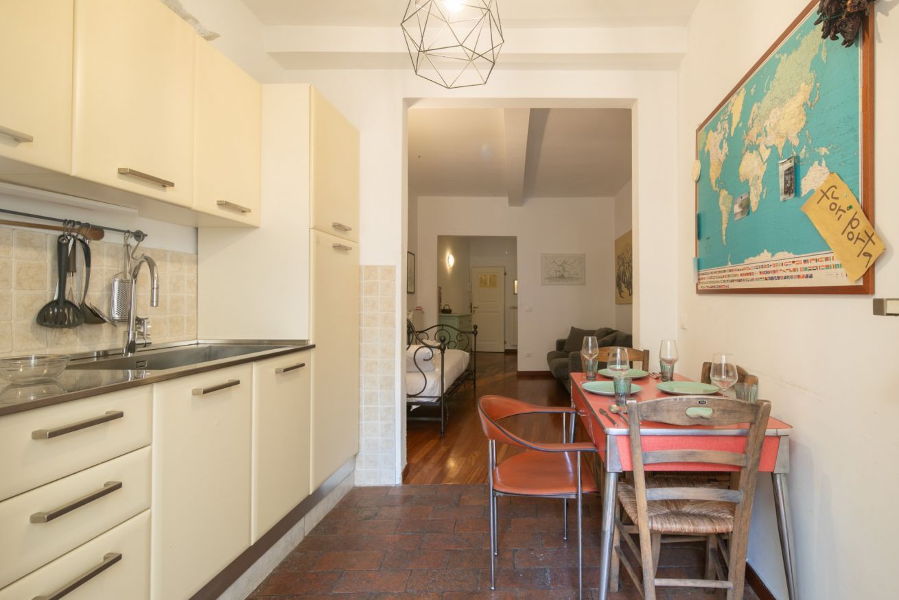 Una cocina compacta con armarios blancos y un salpicadero de azulejos. Se observa un comedor con una mesa y sillas rojas.