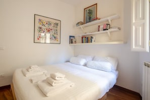 A neatly made bed with folded white towels, a floral painting, and bookshelves with books and a vase. - San Niccolò Elegant Apartment (Florence)
