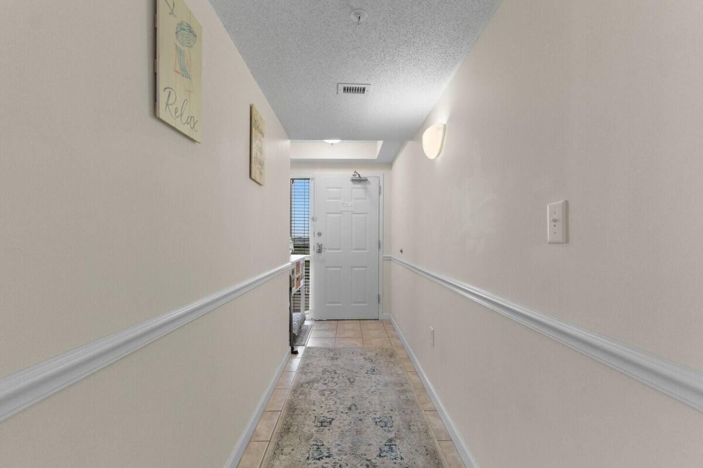 A narrow hallway with a patterned rug, a white door at the end, and a light switch on the wall.