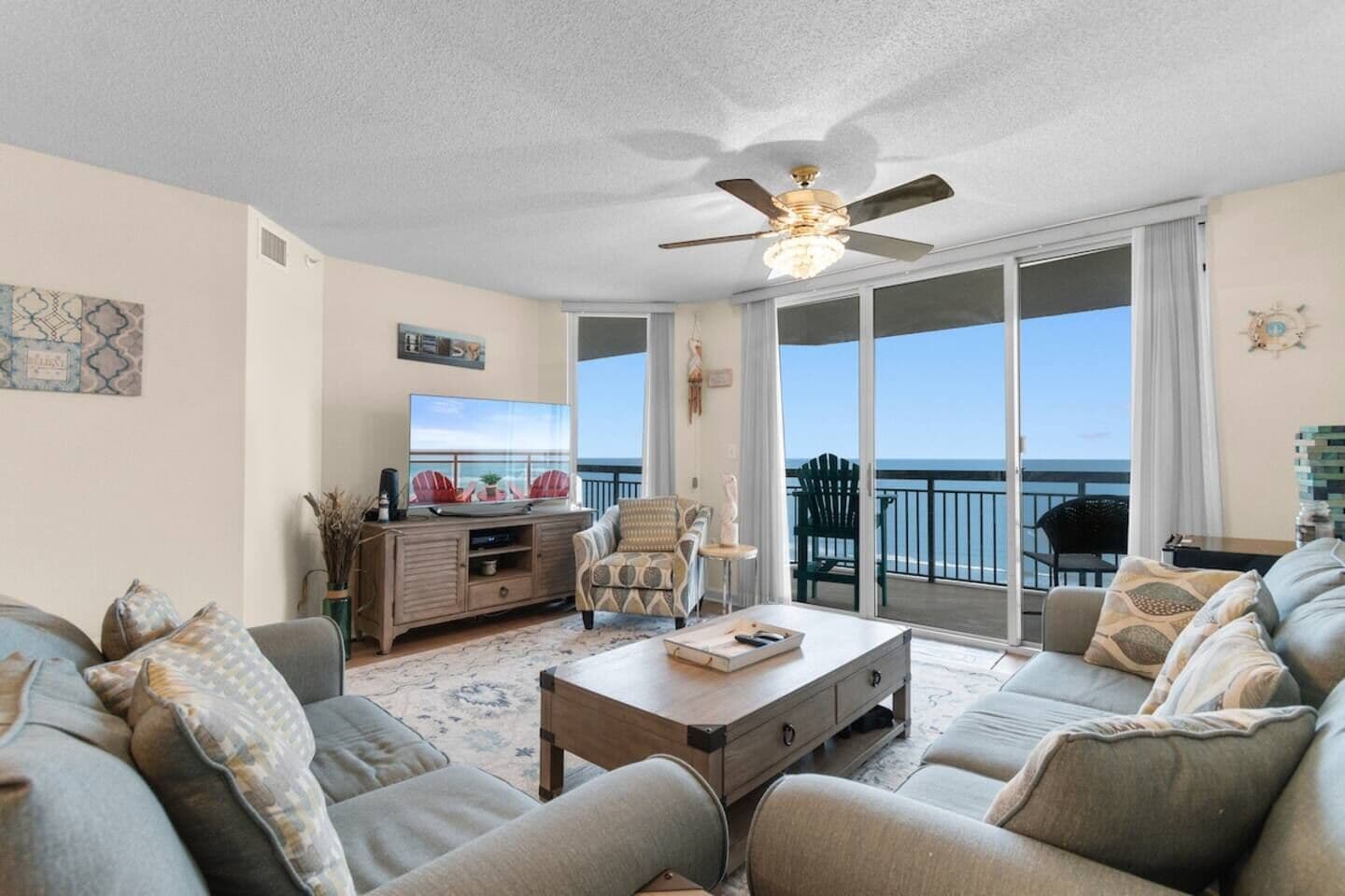 A living room with a sofa, a coffee table, a TV, and a balcony with a view of the ocean.