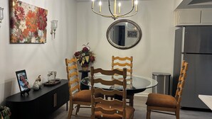 A modern dining area with a round glass table, wooden chairs, a chandelier, a floral painting, and a refrigerator in the background. - This home has been rented annually and not available on VRBO (Cape Coral)