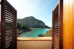 A balcony with wooden shutters, a view of a coastal area with a large rock formation, and a clear blue sea.