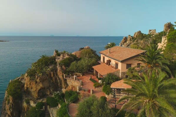 A coastal property with a stone pathway leading to a building with a tiled roof, surrounded by lush greenery and overlooking the sea.