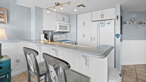 A kitchen with white cabinets, a microwave, a stove, and a refrigerator. There are two bar stools and a small table with a lamp. - 🐬GS Plantation Resort Conference Center 1254 | Resort Amenities+Walk to Beach (Gulf Shores)
