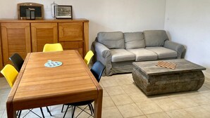 Un salon avec une table à manger en bois, des chaises jaunes, un canapé gris et une table basse en pierre sur laquelle est posé un jeu d’échecs.