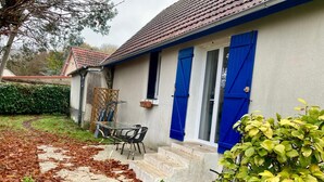 Une maison à l’extérieur blanc, avec des volets bleus et une porte en verre. On y trouve une terrasse avec une table et des chaises, ainsi qu’un jardin avec des plantes vertes et un arbre.